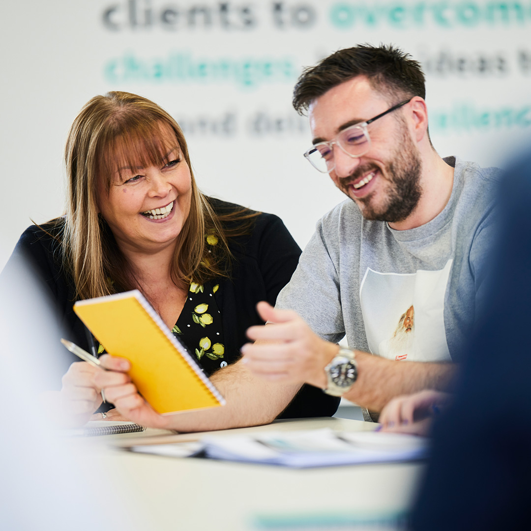 a smiling man and woman sat at a desk looking at a yellow notebook.
