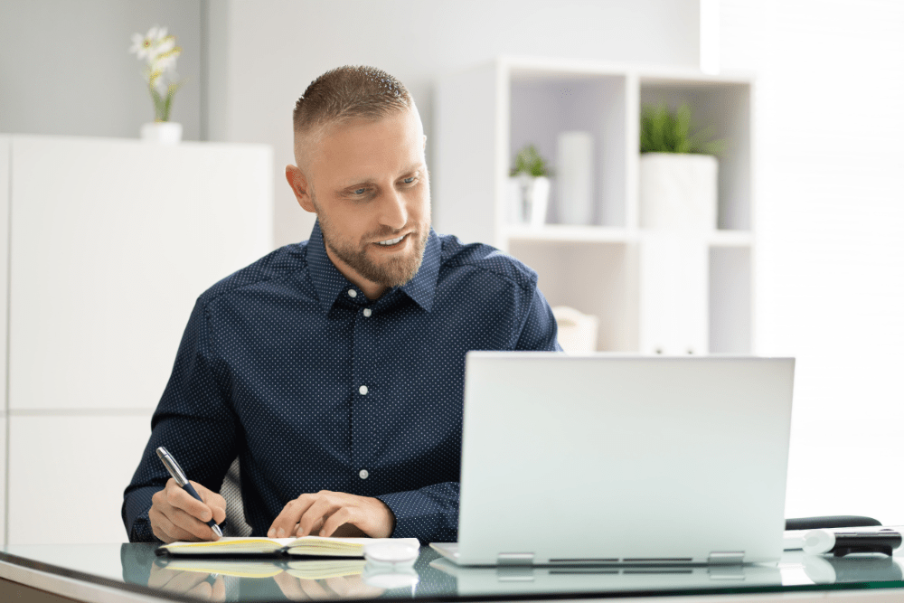 A man completing online training on a laptop whilst taking notes.